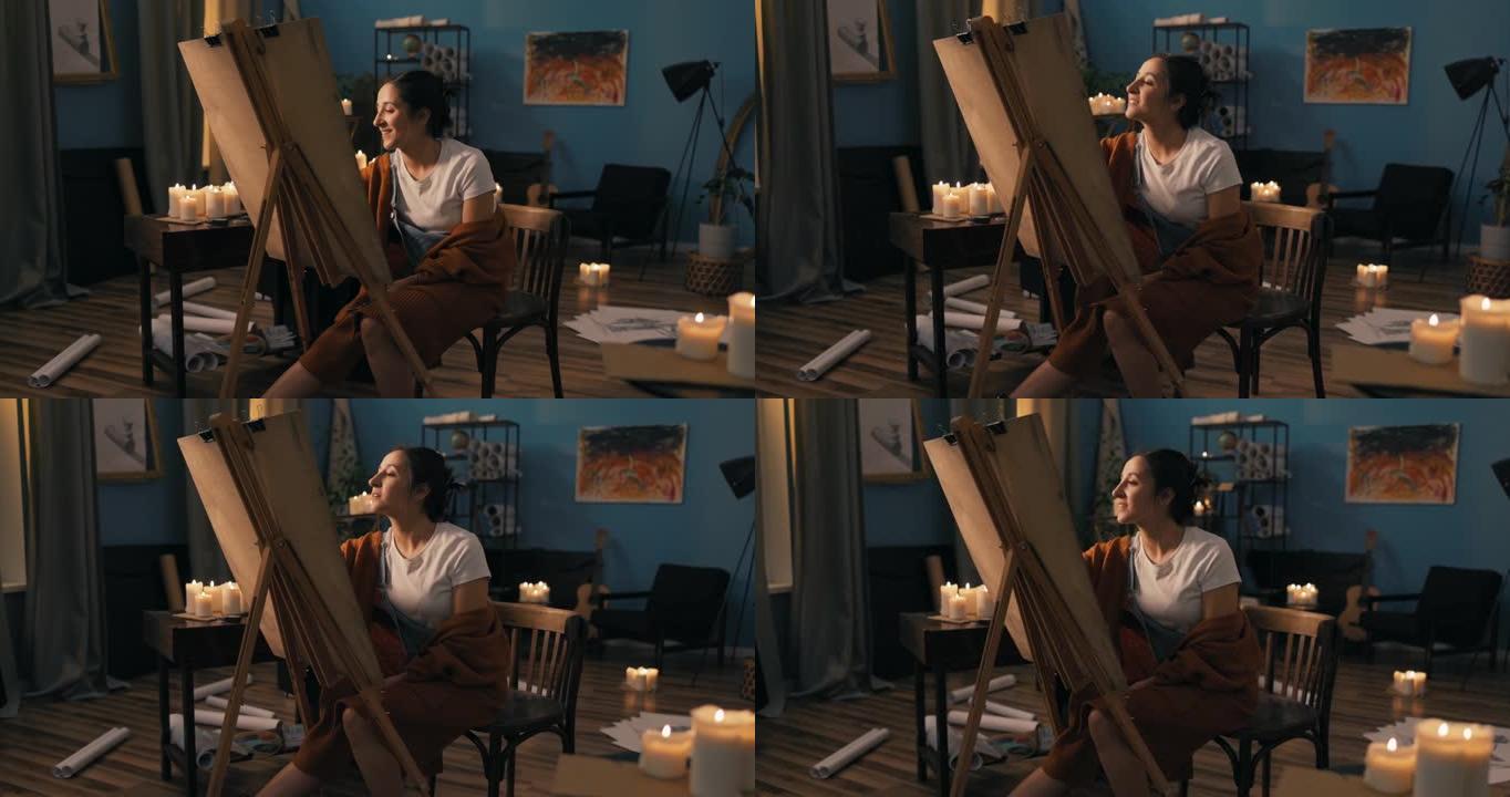 艺术系学生坐在远处教室的家中为大学画项目美丽的女人拿着画布站在画架上分析完成的草图最后的触摸微笑看着