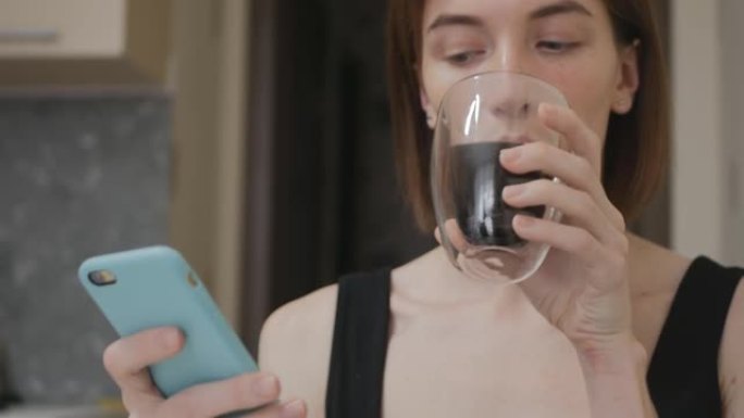 一位年轻漂亮的白种人棕色头发的女人正在绿松石手机中输入一条消息，并用双层玻璃杯喝一杯饮料。早餐和美好
