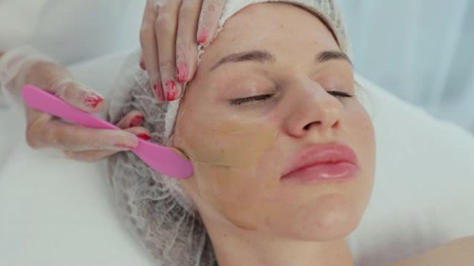 特写美容师用刷子将面膜涂在躺在水疗中心按摩台上的美女脸上。