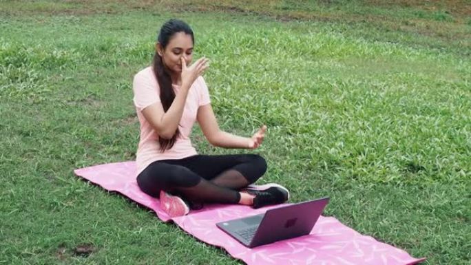 漂亮的印度女人穿着运动服坐在莲花位置的垫子上打开笔记本电脑放松音乐缓解压力做冥想练习。