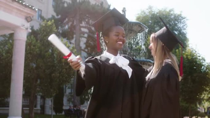 一个美丽的多种族的两个学生女士们在毕业典礼上互相拥抱，他们穿着毕业套装和帽子
