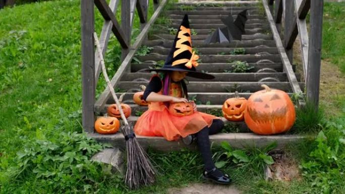 一个穿着女巫狂欢节服装的女孩，戴着扫帚和一顶巨大的黑帽子，在旧的木制台阶上玩耍