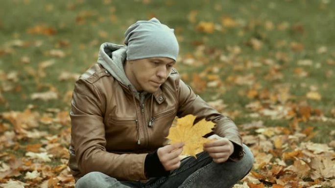 一个戴着帽子的年轻悲伤的人坐在落叶之间，看着一片泛黄的枫叶