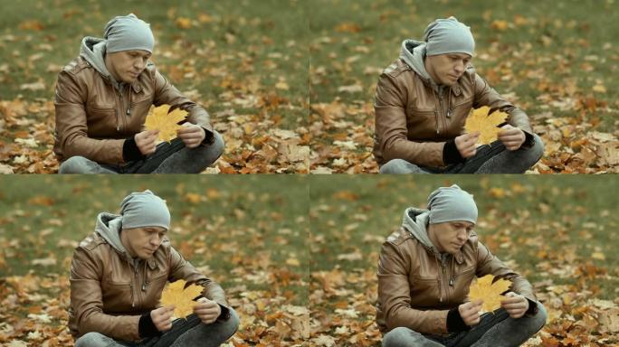 一个戴着帽子的年轻悲伤的人坐在落叶之间，看着一片泛黄的枫叶