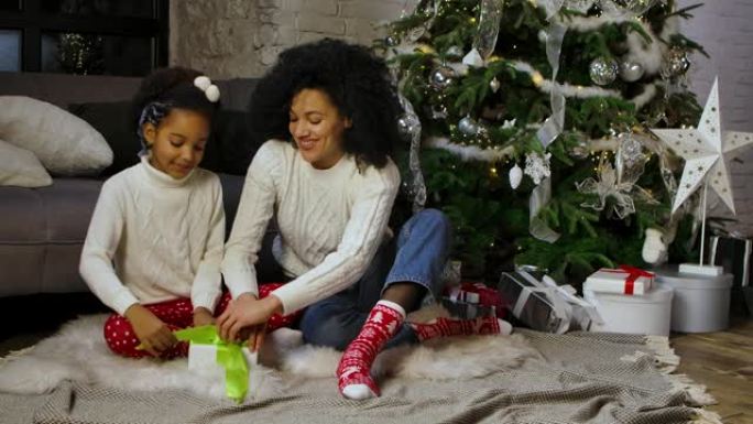 非裔美国妈妈和她的小女儿在礼品盒上系蝴蝶结的肖像。女人和女孩坐在沙发和装饰圣诞树附近。慢动作
