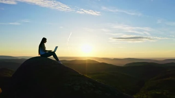 日落时，一名年轻的自由职业者在坐在落基山顶上的笔记本电脑上工作的剪影的空中照片。