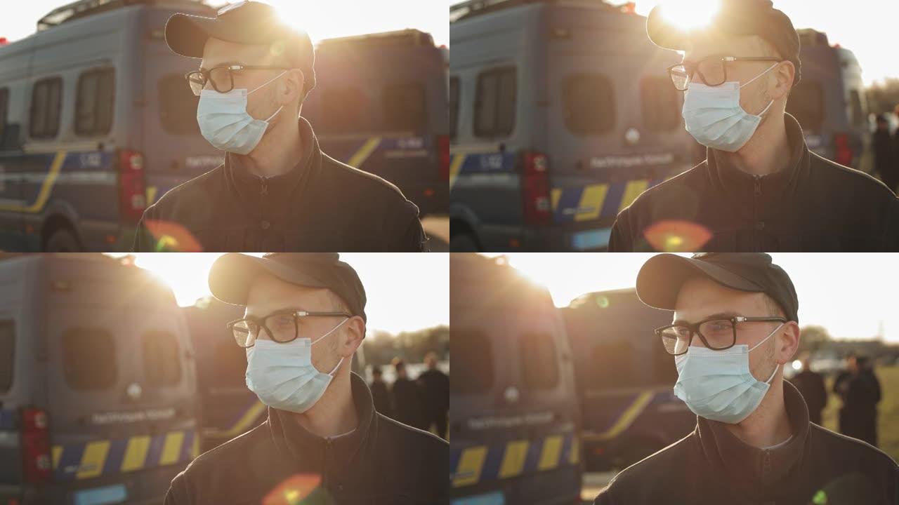 一名身穿警服的男子站在两辆警车附近。他戴着防护面具。他戴着眼镜。阳光明媚。4K