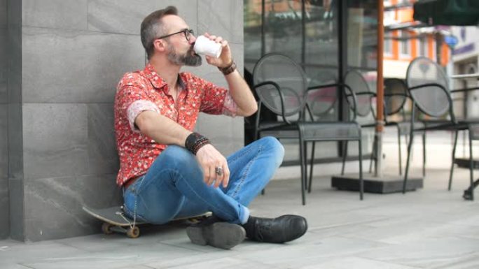 时髦的男人坐在滑板上，在城市喝咖啡