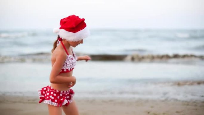 穿着红色泳衣和圣诞老人帽子的5岁女孩在海滩上快乐，跳舞，跳跃和奔跑快乐。圣诞节和新年冬季温暖国家之旅