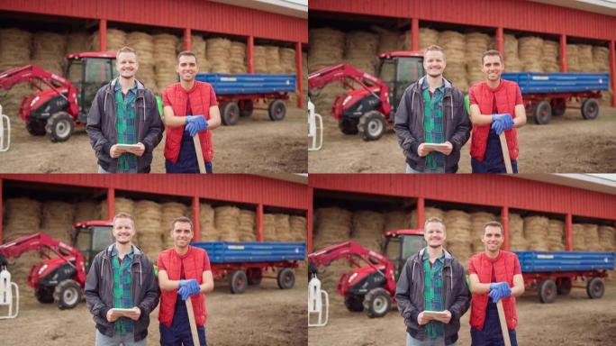 带记事本的农民肖像和带黑桃的农场工人看着相机，微笑着站在户外干草仓库旁边，背景是联合收割机和箱子拖车