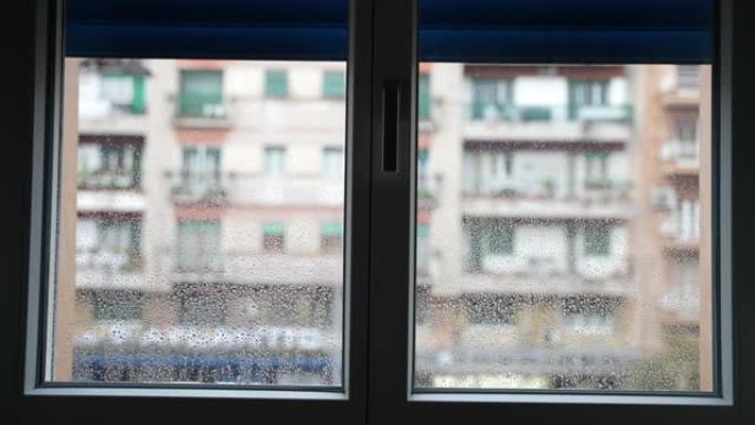 雷雨过后，从潮湿的窗户表面的房间内可以看到，雨天模糊的住宅楼背景上的蓝色百叶窗关闭的窗户。沉重时的悲