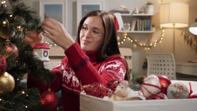 装饰圣诞树。穿着红色毛衣的快乐微笑的女人坐在房间的地板上，把圣诞玩具放在树上。慢动作
