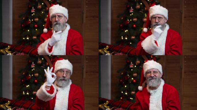 看着相机的圣诞老人肖像专心地听着，并做出一个手势。穿着红色西装和帽子的老人在带有玩具和灯光的圣诞树的