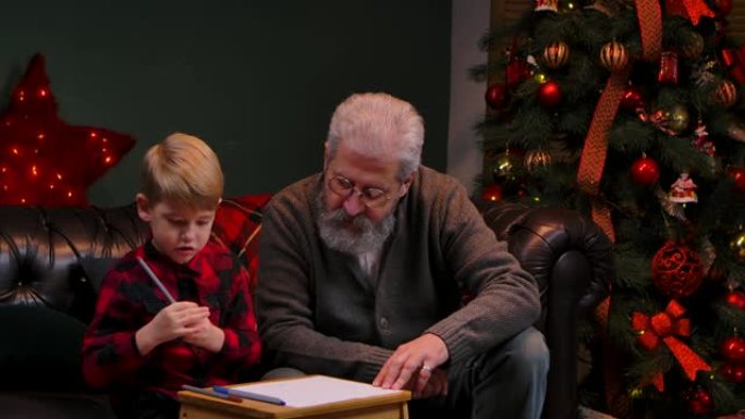 小男孩用毡尖笔画了一幅喜庆的画。祖父和孙子坐在靠近发光圣诞树的装饰房间的沙发上。家庭度假概念。特写。