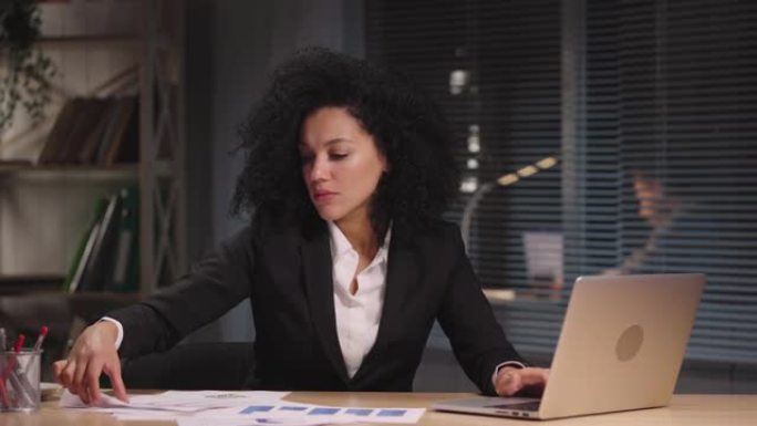非裔美国妇女的肖像正在查看构成报告的图表。女商人在工作场所摆姿势，坐在办公室内部的桌子旁。慢动作准备
