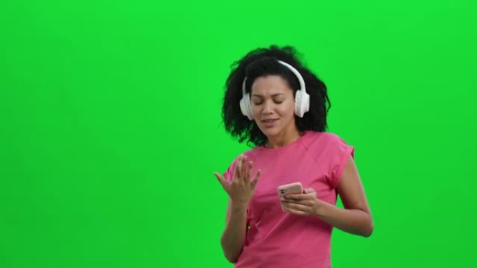 使用智能手机在白色大耳机中跳舞和欣赏音乐的年轻女性非裔美国人的肖像。卷发的黑人妇女在绿色屏幕上摆姿势