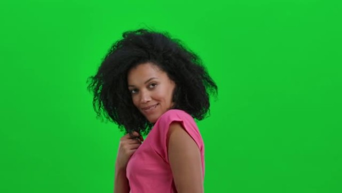 年轻女性非洲裔美国人的肖像在镜头前轻柔地微笑着眨眨眼。卷发的黑人妇女在工作室的绿色屏幕上摆姿势。特写