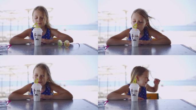 一个开朗，微笑的女孩在暑假期间在海滩上喝清爽的鸡尾酒。眼镜从风中从桌子上掉下来。