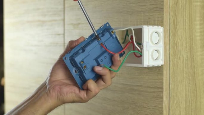 电工正在用螺丝刀将电线拧紧到木墙上的电气插头上。