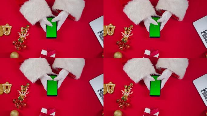 顶视图圣诞老人手使用智能手机与绿色屏幕色度键由红色新年装饰桌。圣诞老人在屏幕上点击观看内容，进行在线