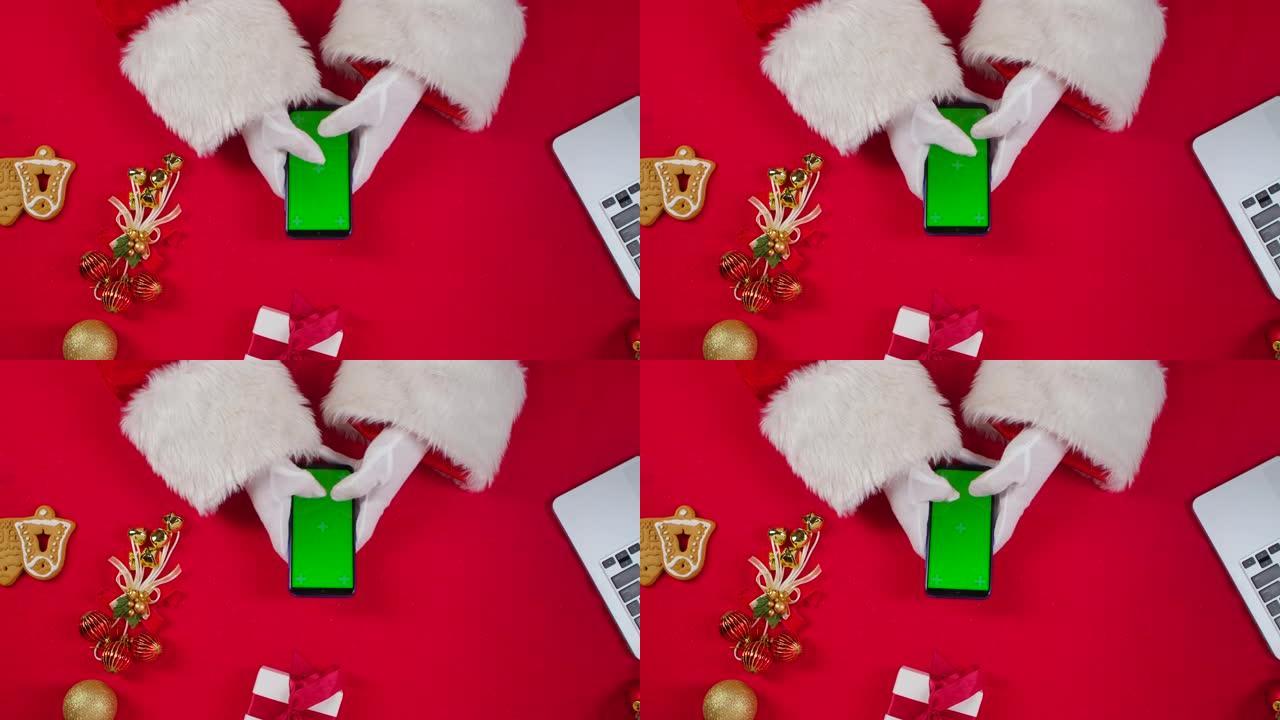 顶视图圣诞老人手使用智能手机与绿色屏幕色度键由红色新年装饰桌。圣诞老人在屏幕上点击观看内容，进行在线