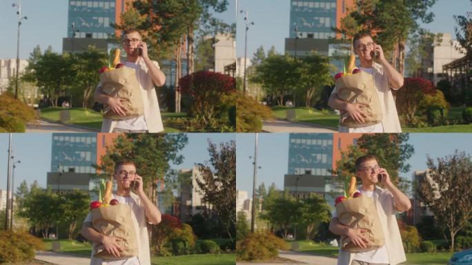 在镜头前，年轻的男人在商店后英俊地走在街上，拿着一个装满蔬菜和水果的生态袋，用智能手机说话