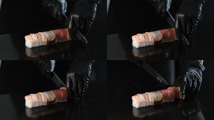 寿司制造商将面包卷切成一块石板。戴着黑手套的厨师正在用刀切卷。