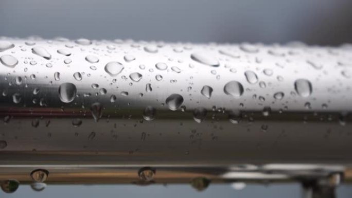 在模糊的海洋背景下，不锈钢扶手上的雨滴。特写镜头。雨露中的水滴聚集在闪亮的钢铬杆手轨栏杆上