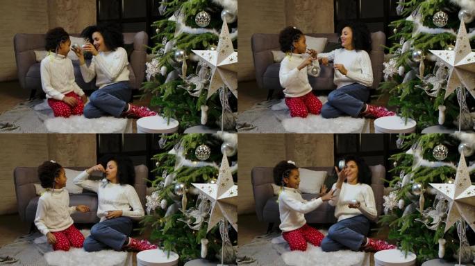 快乐的非洲裔美国妈妈和小女儿的肖像用节日玩具装饰圣诞树，坐在阁楼风格的家庭房间背景下玩得开心。慢动作