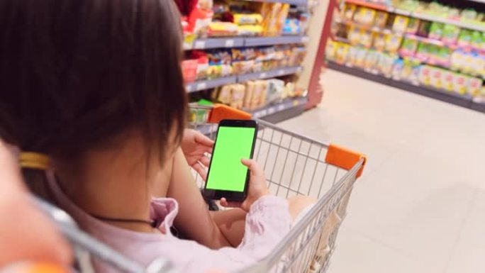 后视图视频，一个女孩坐在杂货店的手推车上，双腿交叉，双手拿着智能手机，绿屏，母亲推着手推车在商店的货