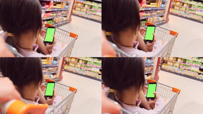 后视图视频，一个女孩坐在杂货店的手推车上，双腿交叉，双手拿着智能手机，绿屏，母亲推着手推车在商店的货