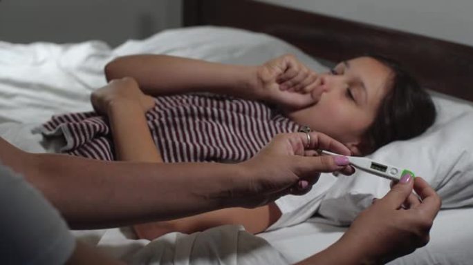 9岁小女孩的特写镜头感冒躺在床上患流感。小孩咳嗽和打喷嚏，母亲的手测量她的高温。4K