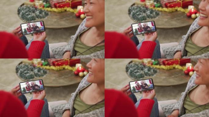 微笑的亚洲夫妇使用智能手机进行圣诞节视频通话，屏幕上戴着圣诞老人帽子的夫妇