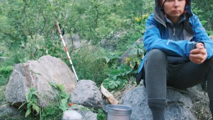 一个女孩坐在石头上，在树林里徒步旅行时，等待锅在燃烧器上煮沸。