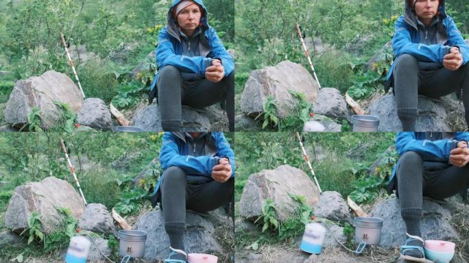 一个女孩坐在石头上，在树林里徒步旅行时，等待锅在燃烧器上煮沸。