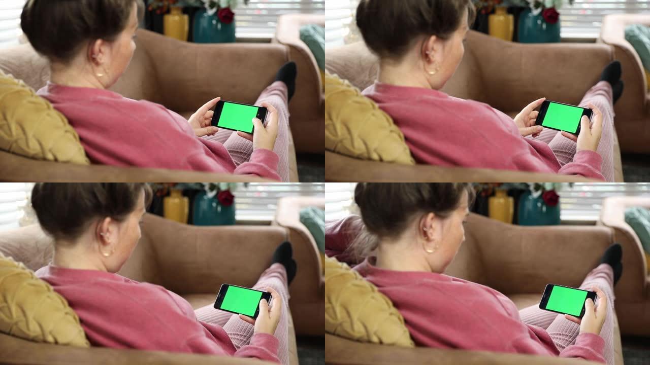 在家中的年轻女子躺在沙发上，在水平景观模式下使用绿色模拟屏幕智能手机。女孩使用手机，浏览互联网，观看