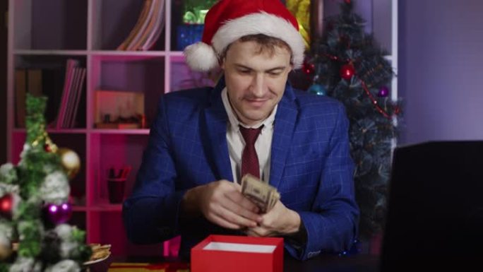 戴着圣诞老人帽子的年轻人打开礼品盒，把钱放在里面，坐在办公室里。新年气氛中的快乐商人包装礼物。预期假