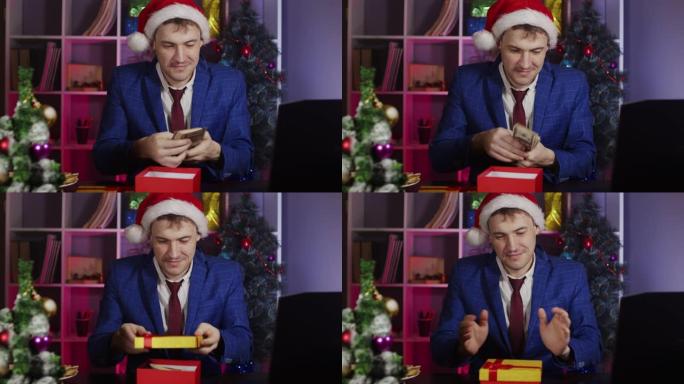 戴着圣诞老人帽子的年轻人打开礼品盒，把钱放在里面，坐在办公室里。新年气氛中的快乐商人包装礼物。预期假