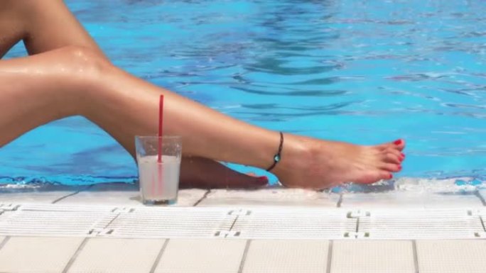 性感的女腿在公共泳池上，用塑料吸管端着一杯鸡尾酒或柠檬汁