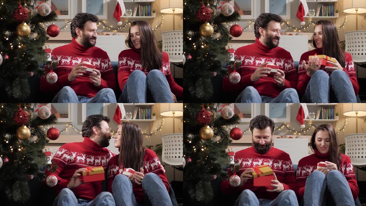 送圣诞礼物。幸福的夫妻男人和女人坐在圣诞树附近的地板上，互相送礼物。慢动作