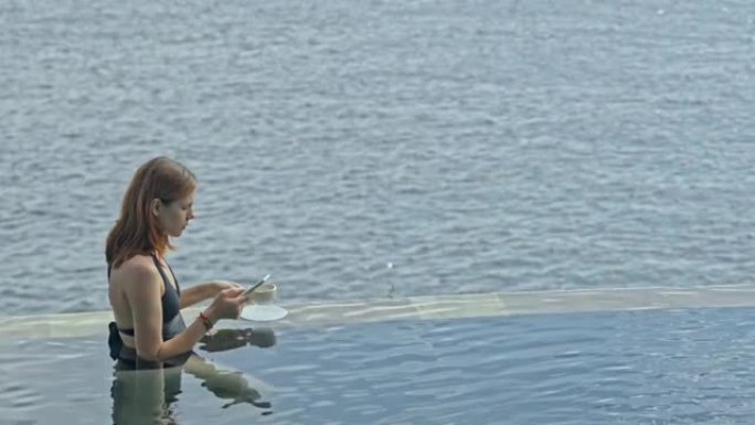 游泳池里的女人使用智能手机。海洋背景上的巴厘岛度假胜地的女性上网