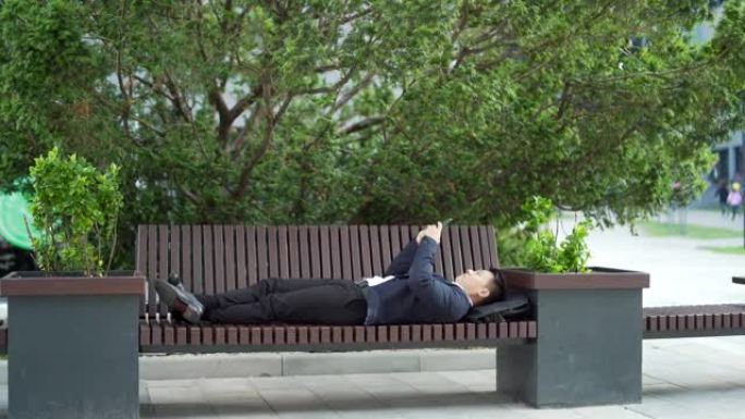 亚洲商人上班族放松躺在长凳上手里拿着手机