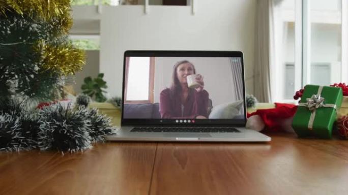 微笑的白人妇女在笔记本电脑上的圣诞节视频通话中喝咖啡