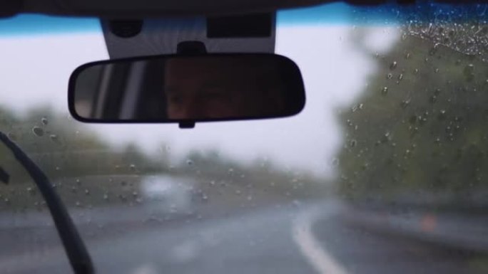 中年男子在驾驶汽车内部的镜子中的反射，在大雨中，驾驶员在公路旅行中握着方向盘并与无法识别的乘客交谈的