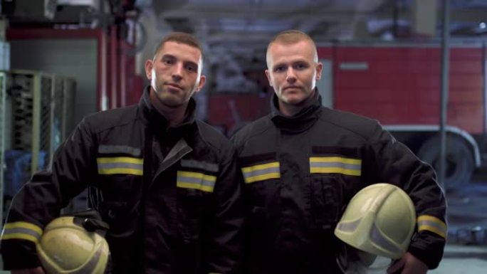 两名消防员站在一起，身穿制服，手持黄色防护头盔。背景中的消防车。摄像机向左移动。