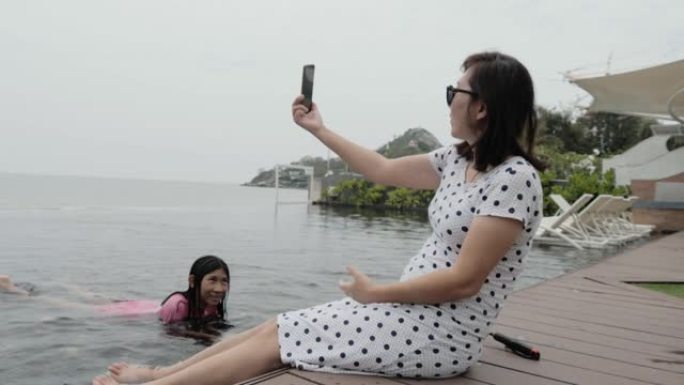 快乐的亚洲妈妈女人在游泳池边放松，在家庭度假期间用智能手机自拍照片，生活方式理念。