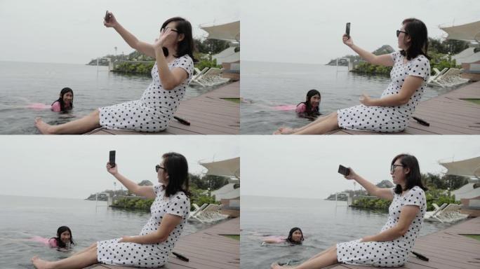 快乐的亚洲妈妈女人在游泳池边放松，在家庭度假期间用智能手机自拍照片，生活方式理念。