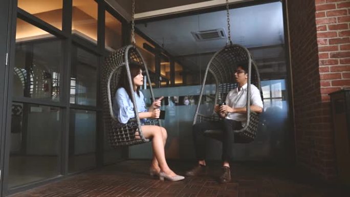 年轻的亚洲女性坐在共同工作空间的藤制吊椅上，与同事一起享受咖啡，谈论商业策略
