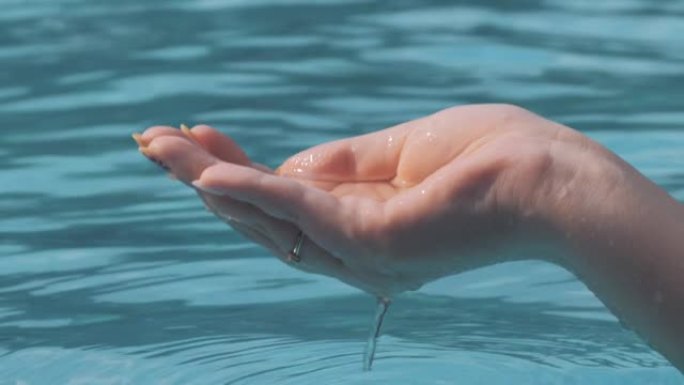 女性手玩水的特写镜头，在阳光明媚的日子用手指抓住并倒入水池。人类手掌舀水，水滴落在水面