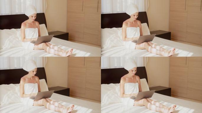 迷人的高加索女人裹着浴巾，用现代笔记本电脑在酒店房间放松。漂亮的女士躺在床上上网。旅行和技术的概念。